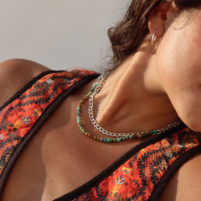 Close-up of a dark-haired woman in a patterned orange top wearing small silver hoop earrings and two layered necklaces: a chunky silver link chain paired with a bead strand of turquoise and earthy brown stones in natural sunlight.
