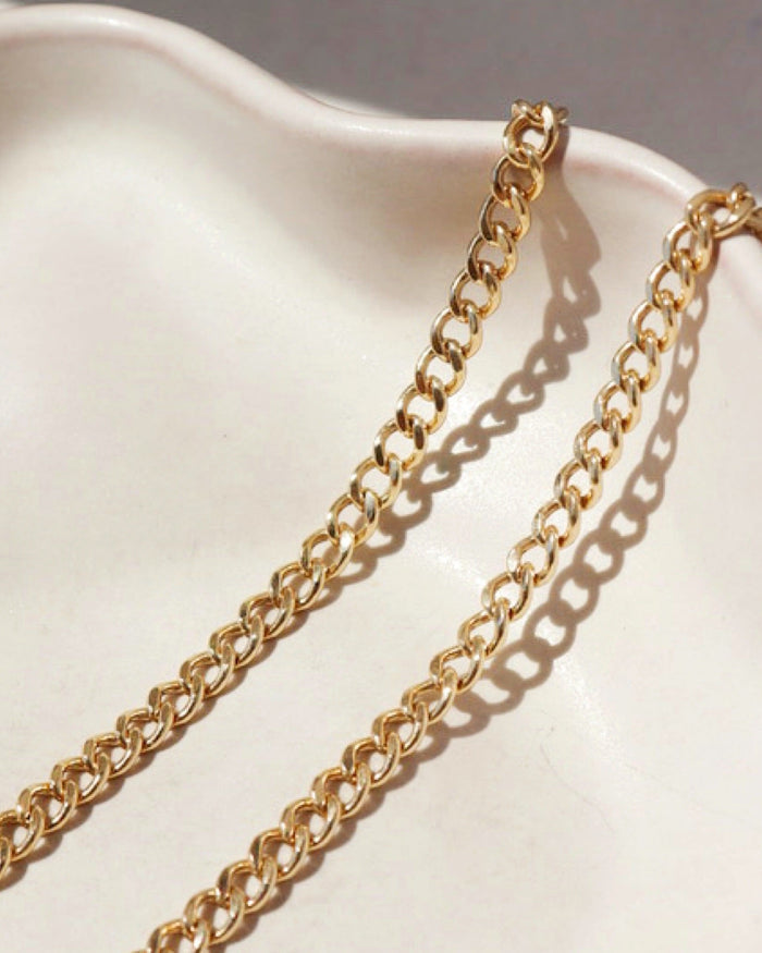 Extreme macro close-up of a gold curb chain resting on a smooth, off-white ceramic dish under direct sunlight.