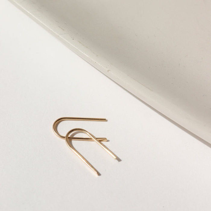 14k solid gold wire formed into arch shaped earrings with one hammered end, photographed on a white table next to a white dish