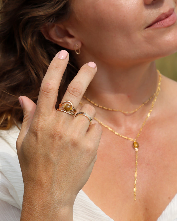 Close-up of a woman wearing layered gold necklaces, Tiger’s Eye rings, and a small gold hoop earring in warm sunlight.