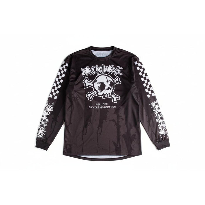 A man wearing the Backbone 2026 Gnar Drip Jersey: a black, lightweight, breathable top with a skull and crossbones graphic, checkerboard sleeves, and "Backbone" plus "Real Deal Bicycle Motocross" printed on it.