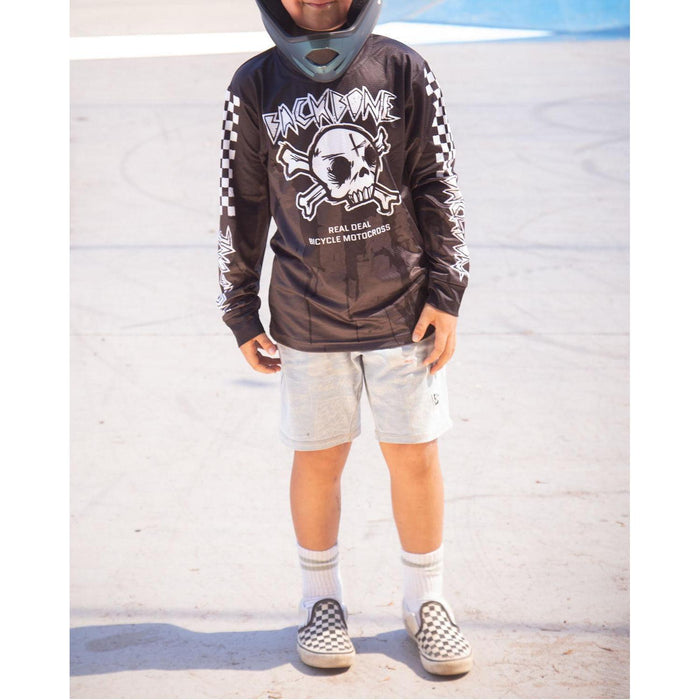 A child in a helmet, Backbone 2026 Gnar Drip Jersey with skull graphic, light shorts, white socks, and checkered slip-on shoes stands on a concrete surface.