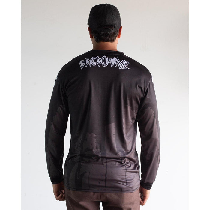 A person faces away, wearing the black Backbone 2026 Gnar Drip Jersey and black cap; bold white "BACKBONE" lettering stands out on the upper back.