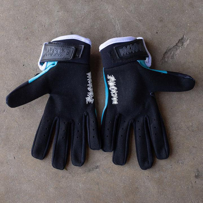 A pair of Backbone Gnar Drip Adult Gloves made from lightweight spandex, in black and teal with Velcro straps and white stitching, lies on a concrete surface.