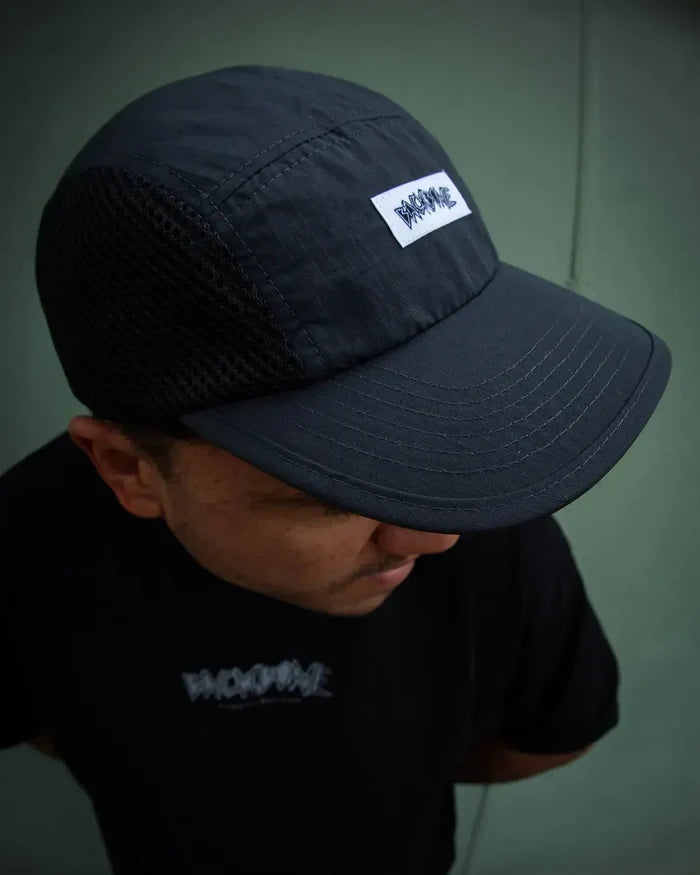 Wearing a black Backbone Ride Club Hat featuring a “BADWAVE” patch, along with a matching black “BADWAVE” t-shirt, the person stands against a plain greenish wall.