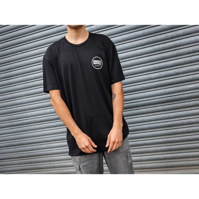 Clad in a black BSD Fully Roasted T-Shirt and gray cargo shorts, the person stands against a corrugated metal backdrop, holding a double shot of coffee for an extra kick.