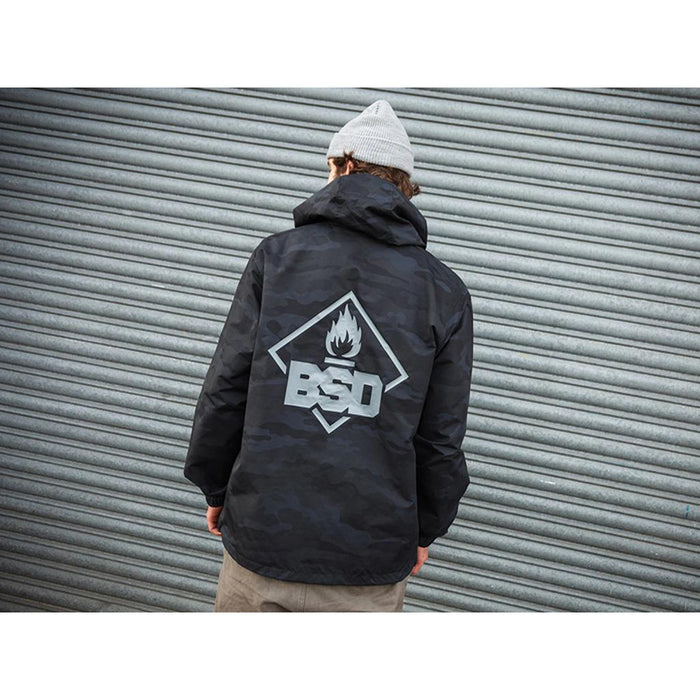 A person wearing the BSD Roastin Jacket, showcasing a black camo with a flame logo and "BSD" on the back, confidently poses in front of a corrugated metal wall while sporting a gray beanie.