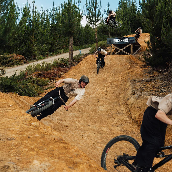 Four cyclists, each sporting the Buzzed Riders Split Tee, ride mountain bikes on a dirt trail with jumps beneath towering pine trees. In the background, a ramp marked "RockShox" hints at high-flying action—making this vibrant scene a true conversation starter.