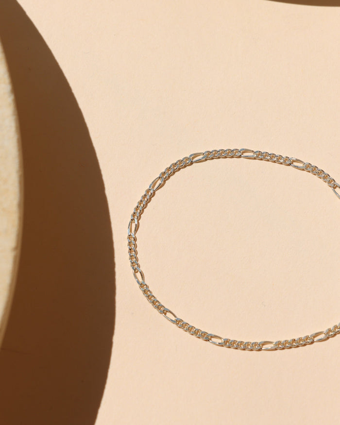 Minimalist sterling silver Figaro chain bracelet featuring alternating short round links and elongated oval links, laid flat on a peach-beige surface with geometric natural light shadows.