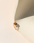 A gold ring with a round brown tiger's eye stone, resting on a textured beige ceramic surface under bright sunlight.
