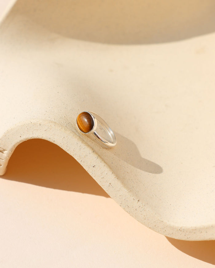 A silver ring with a polished oval cabochon of tiger's eye stone, displayed on a textured beige ceramic surface under warm sunlight.