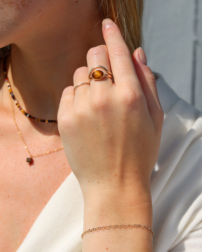 A close-up lifestyle shot of a woman's hand wearing a rose gold ring with a polished amber stone, along with other delicate gold jewelry.