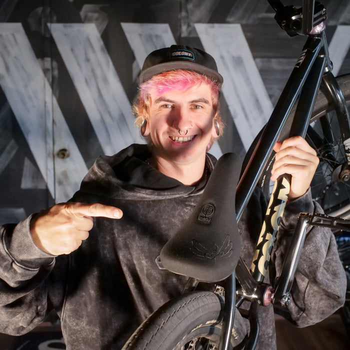 A person with dyed hair and piercings smiles and points at the Colony Sigil Pivotal Seat (Sean Gardner Signature) on a BMX bike indoors.