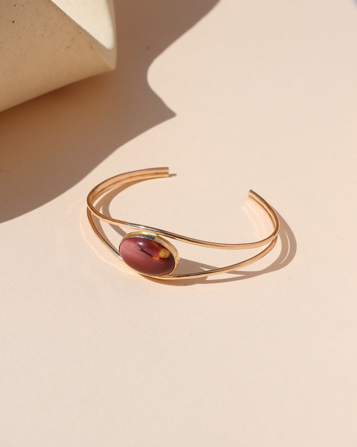Gold open cuff bracelet with an oval red and burgundy stone centerpiece, photographed in warm sunlight on a cream background.