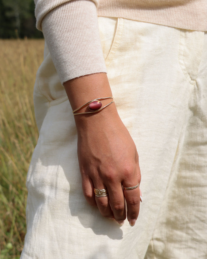 Gold open cuff bracelet with an oval red stone centerpiece, worn on the wrist and styled with gold rings and a cream outfit outdoors.