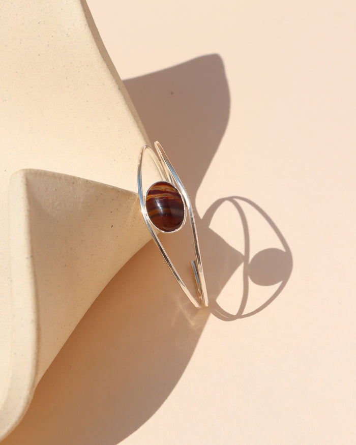 Silver open cuff bracelet featuring an oval banded stone in deep red and brown tones, displayed against a cream ceramic form in warm sunlight.