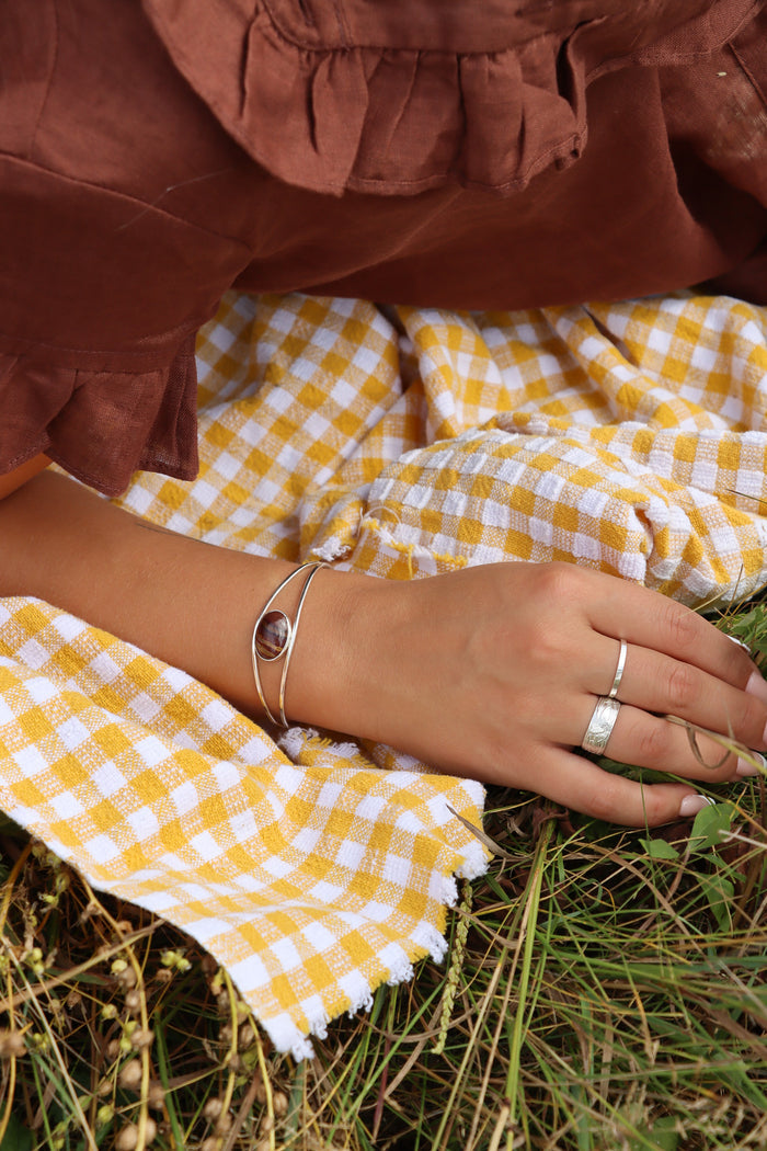 Silver open cuff bracelet with an oval banded red and brown stone centerpiece, worn on the wrist and styled with silver rings against yellow gingham fabric.