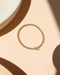 Classic gold curb link chain bracelet featuring interlocking flattened links and a spring ring clasp, laid flat on a peach-beige surface near a ceramic dish with warm natural light shadows.