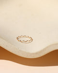 Minimalist gold ring with a gentle squiggly wave pattern, displayed inside a cream-colored textured ceramic dish with soft natural sunlight and warm shadows.