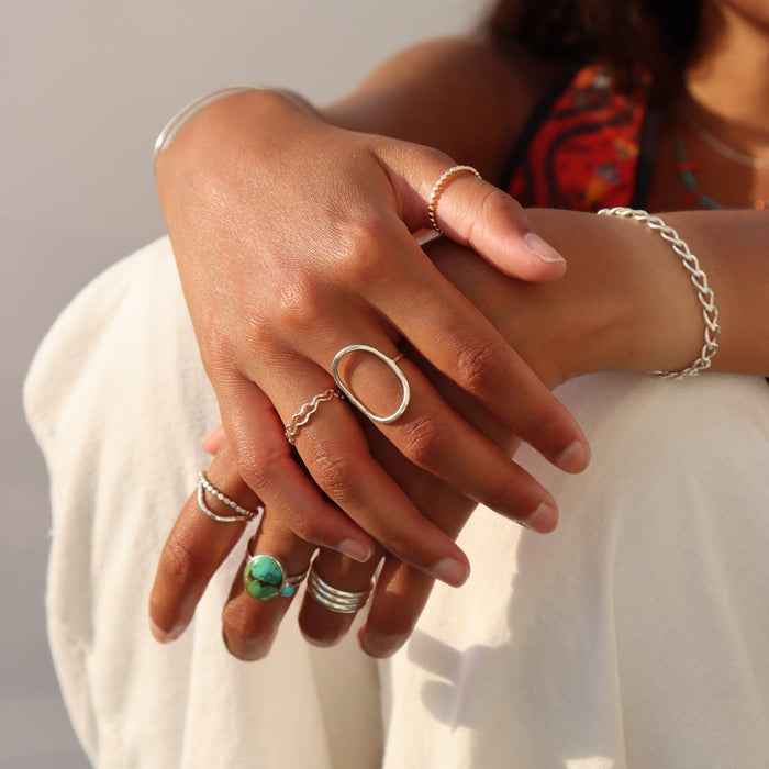 Lifestyle image of layered sterling silver rings, including a bold oval statement ring, natural turquoise ring, beaded stacking rings, and a wavy band paired with a sterling silver chain bracelet in warm natural sunlight.