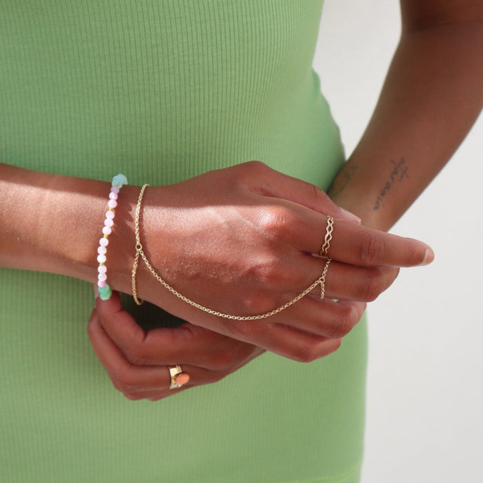 Model wearing two 14k gold fill rings with wave-like textured wire, model is also wearing a hand chain made with a 14k gold fill rolo chain, paired with a bright mixed gemstone bracelet.