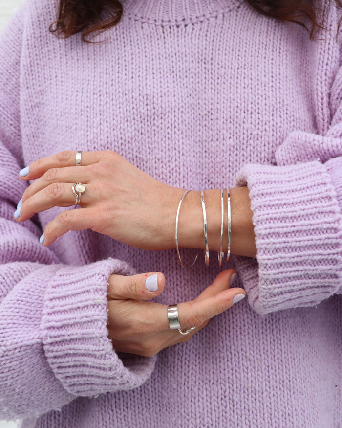 A close-up of sterling silver bangles stacked on a wrist, paired with silver statement rings against a cozy lavender knit turtleneck sweater.