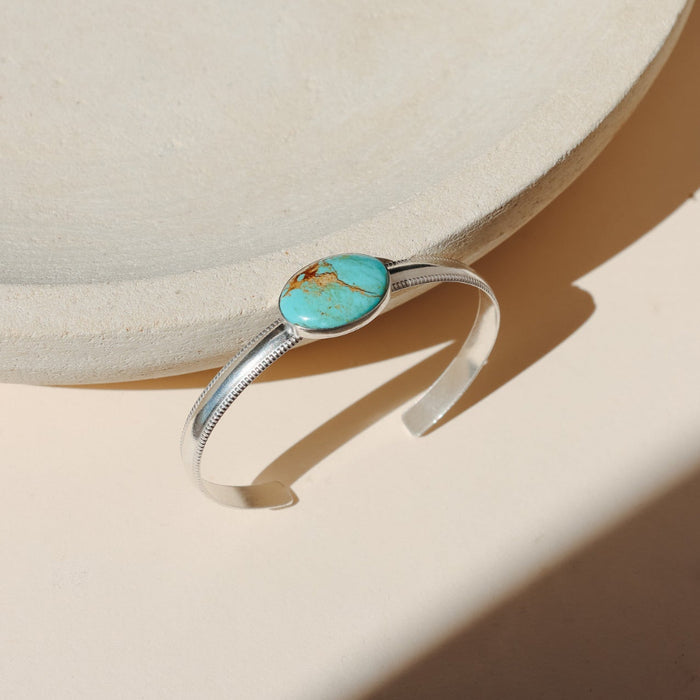 Sterling Silver bezel set turquoise cuff on a thick smooth band with textured edges on a beige background in the sunlight