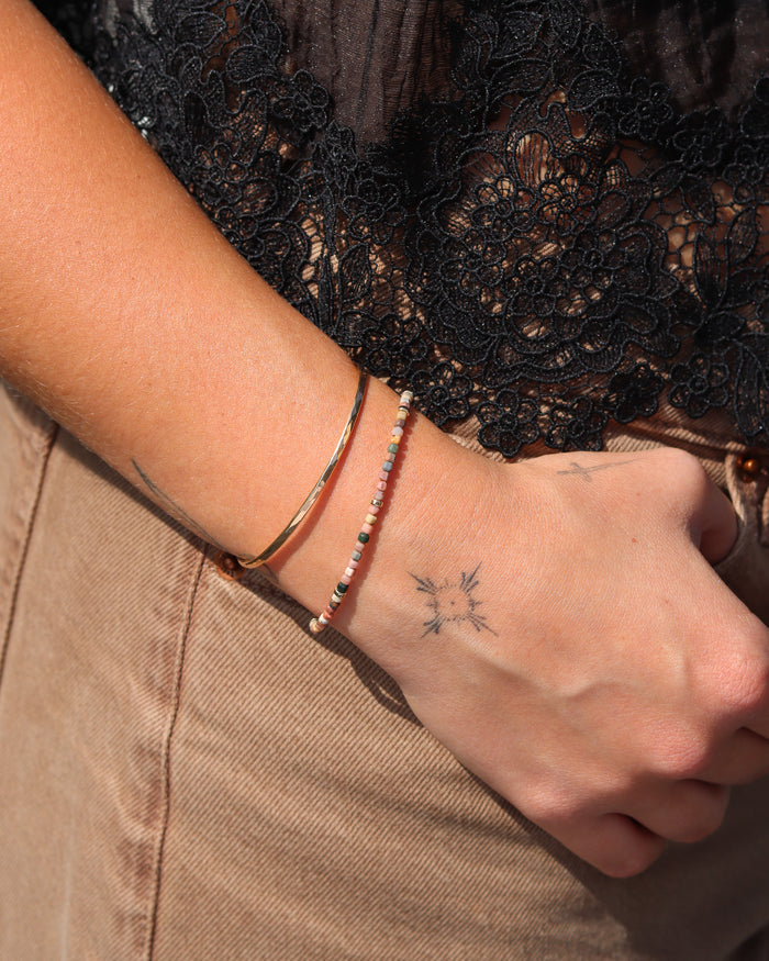 A close-up of a person's wrist wearing two delicate bracelets: a thin gold bangle and a beaded bracelet with pink, white, and dark beads. The person is wearing a black lace top and tan pants.