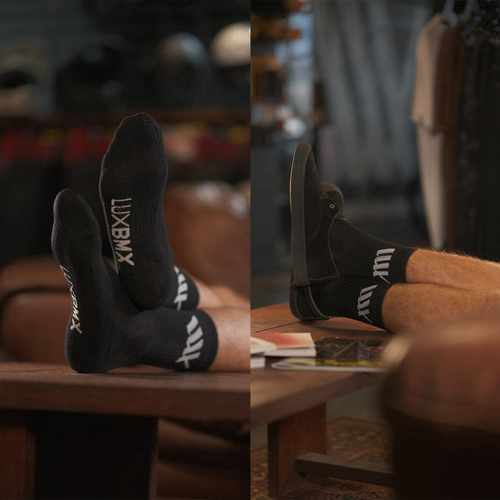Close-up of a person wearing black LUXBMX Crew Socks (3 pack), feet on a table, with indoor furniture and a blurred background.