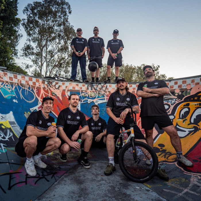 Eight men in matching dark athletic gear, including a standout LUXBMX MMX Soccer Jersey - Plain Back Print, pose in a colorful graffiti-covered skate bowl; some are standing while others sit casually, with one person holding a bike.
