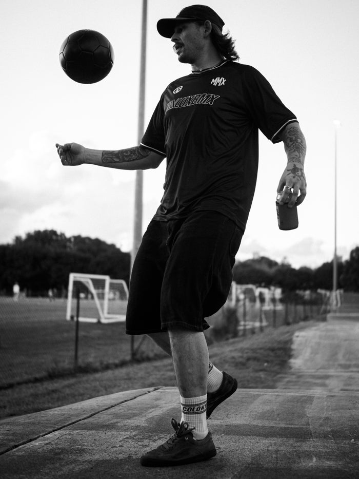 Wearing a LUXBMX MMX Soccer Jersey - Plain Back Print, a man juggles a soccer ball on one foot and holds a drink, enjoying the outdoors near soccer fields.