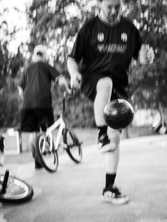 A person in sportswear juggles a soccer ball on one foot while another person stands nearby with a bicycle, wearing a breathable LUXBMX MMX Soccer Jersey - Plain Back Print.