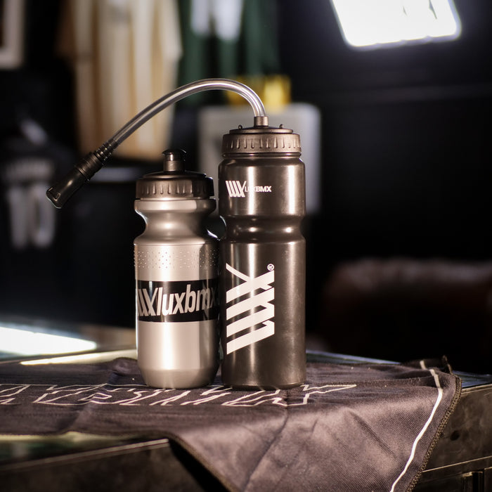 Two LUXBMX Squirt Water Bottles 500ml—one silver, one black—made from BPA-free plastic, with "XXluxbmx" branding, rest on black cloth in a dimly lit indoor setting.
