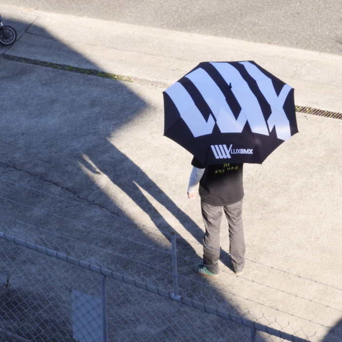 A person stands on concrete holding a LUXBMX Race Track Umbrella with bold white "LUX" lettering, casting a shadow near a chain-link fence.