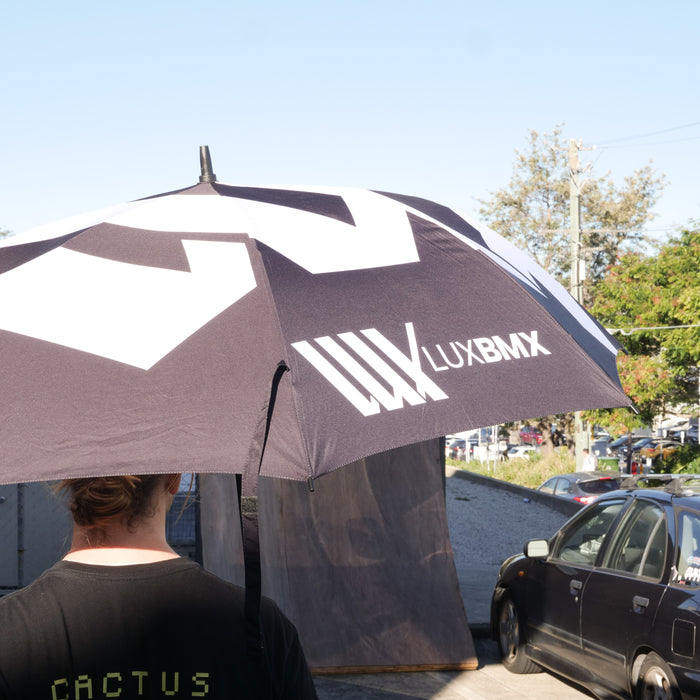 A person holds the LUXBMX Race Track Umbrella with a sturdy carbon pole, standing near parked cars on a sunny day.