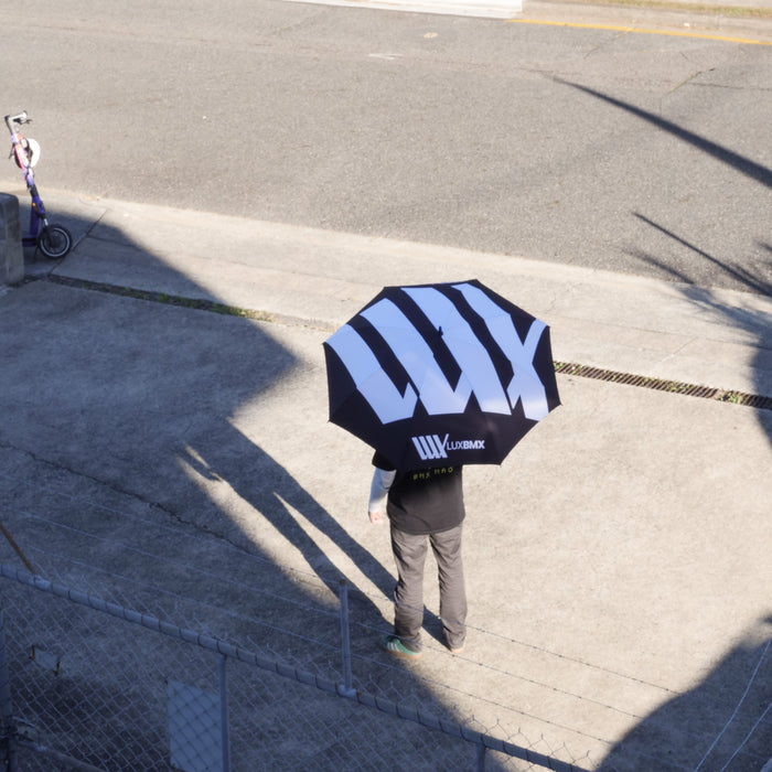 A person stands on a concrete sidewalk holding a LUXBMX Race Track Umbrella with bold white "LUX" lettering; a scooter is parked nearby.