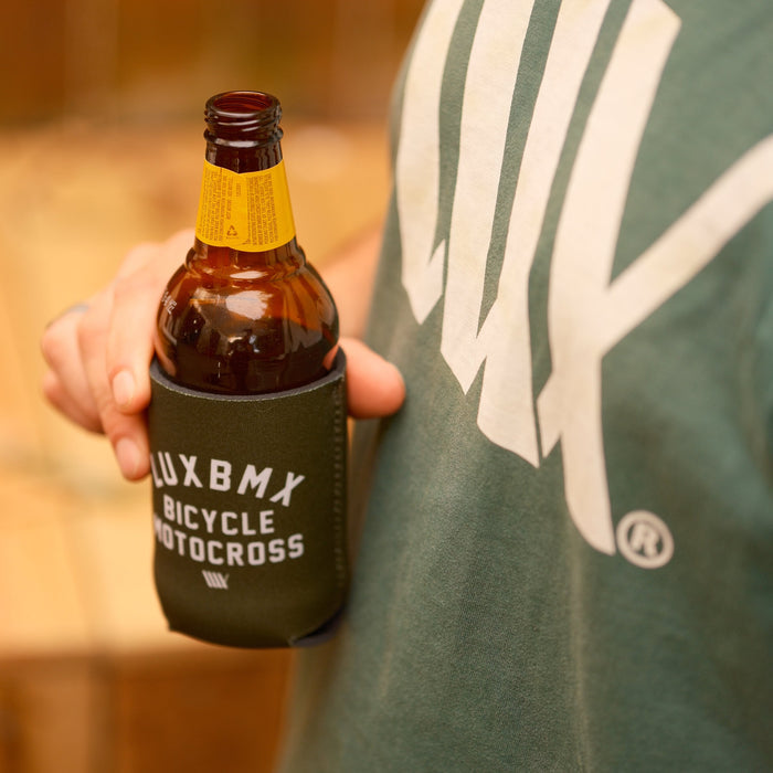 A person holds a brown beer bottle in a green LUXBMX Stubby Cooler / Bicycle Motocross insulator, wearing a green shirt with a large white graphic.