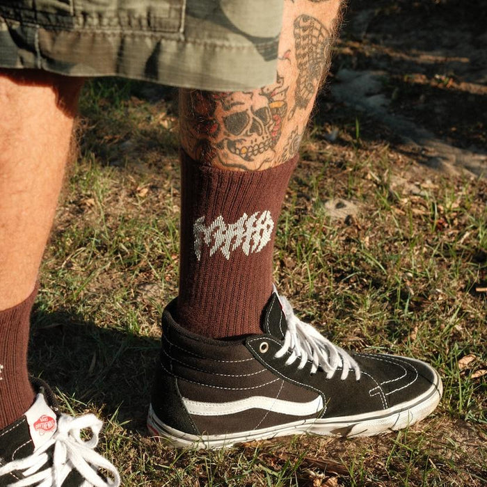 A person stands on grass wearing camouflage shorts, black Vans sneakers, and Mahd Metal Socks—bamboo blend brown riding socks with white text (one size fits most); tattoos are visible on their leg.