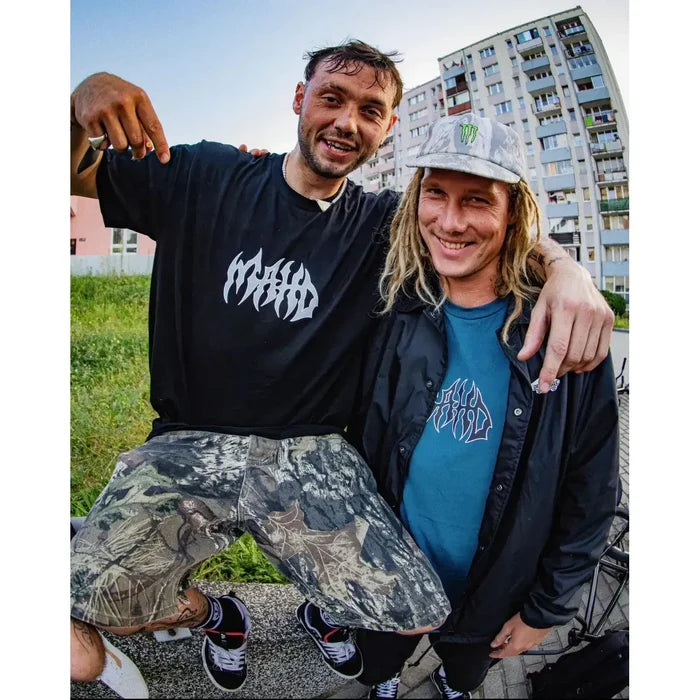 Two men pose outside near apartments; one points to his black Mahd Metal Tee with a bold center logo, while the other, in a gray cap and blue graphic tee with a central design, stands beside him.