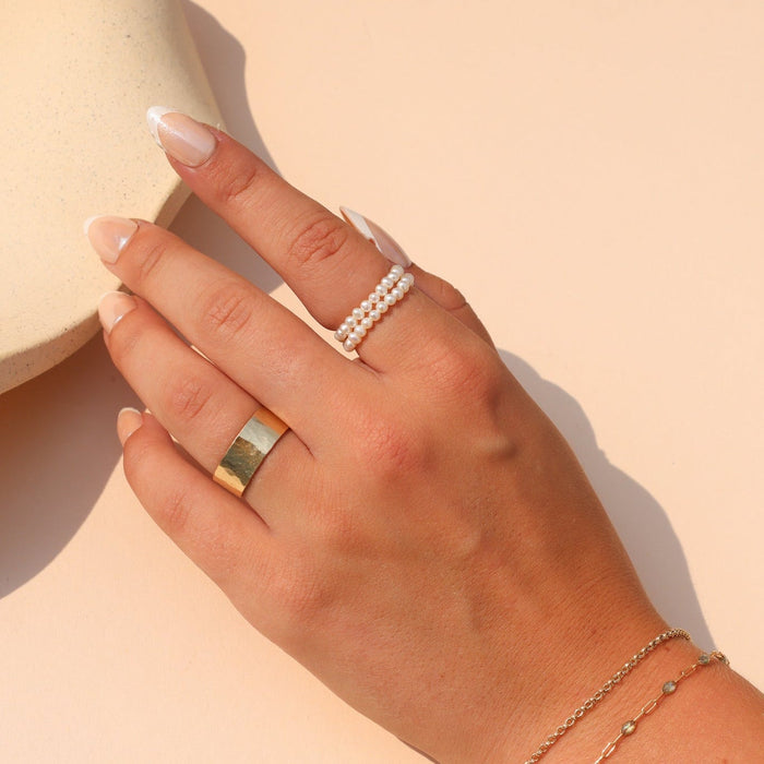 Delicate double-row pearl ring styled with a gold statement ring and layered gold bracelets in warm sunlight.