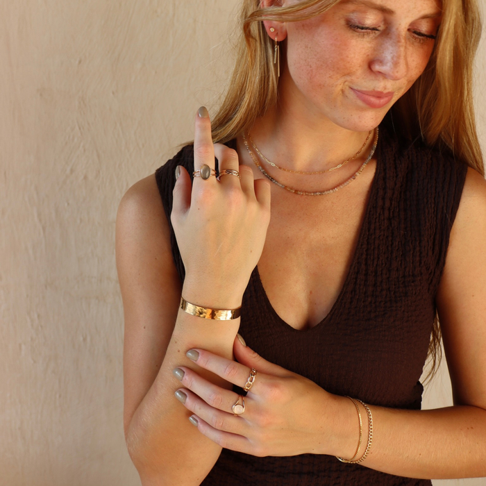 Model wearing the 14k Gold Fill Mirage Cuff, a polished hammered gold cuff bracelet, styled with gold rings, necklaces, and earrings in warm natural light.