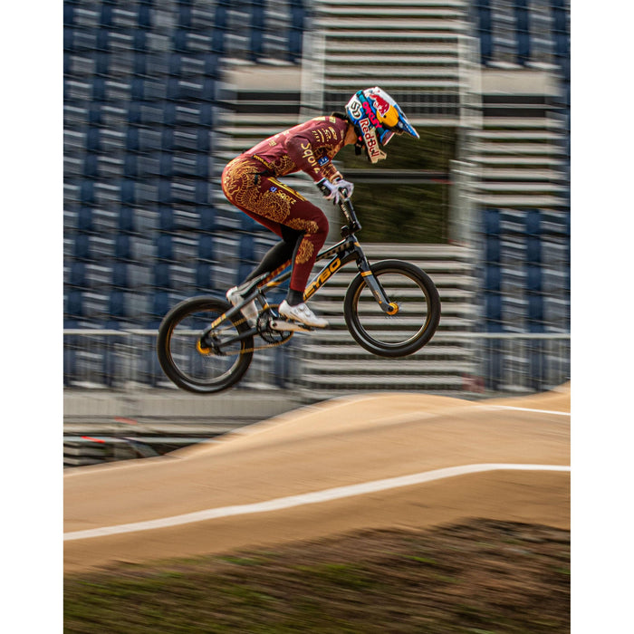 A cyclist in a maroon and gold suit and helmet jumps over a dirt track on a BMX bike fitted with Schwalbe SX-R 20 Inch Tyres, with empty bleachers in the background.