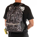 A person is wearing the Staystrong Backpack Chevron V3, featuring a gray and black camouflage design, a front pocket, mesh side pocket, and adjustable shoulder straps. They are also dressed in a black t-shirt with white lettering.