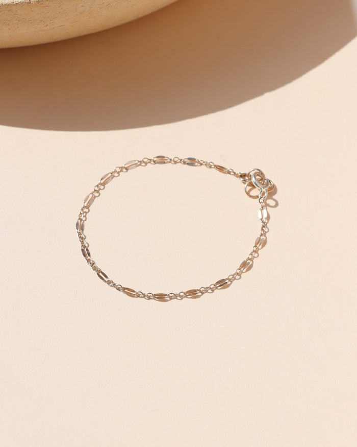 A delicate silver chain bracelet arranged flat in a circular shape on a light beige surface. The fine chain features alternating flattened oval links and a spring ring clasp, set against a warm background with a ceramic tray edge casting soft shadows.