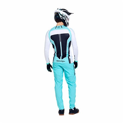 A person wearing turquoise pants, gloves, a helmet, and the TLD 26.1 Sprint Ultra Jersey in Proton White with a black back protector stands facing away from the camera.